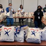 Nine CASA staff members in a large room stand beside tables stacked with CASA care bags, May 2020.