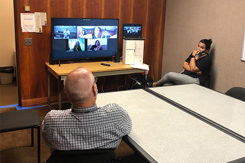 Counselor Stephen Holbrook and Probation Officer Ana Ochoa wearing masks and sitting more than six feet apart in a room watching the 72nd Drug Court Graduation proceedings on a television showing the video feeds of people participating remotely from different locations.