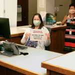 Librarian Marlene Cuenco and State Law Librarian Jenny R.F.F. Silbiger wearing masks and holding up hand sanitizer and the reservations phone number at the Hawaii Supreme Court Law Library reference desk, 08/03/2020.