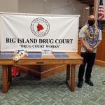 In the courtroom, two graduates stand on the left and right sides of a table holding a Big Island Drug Court banner (“Drug Court Works”), with Hilo Drug Court Judge Henry Nakamoto on the far right, at the program’s 57th graduation ceremony, 08/05/2021.